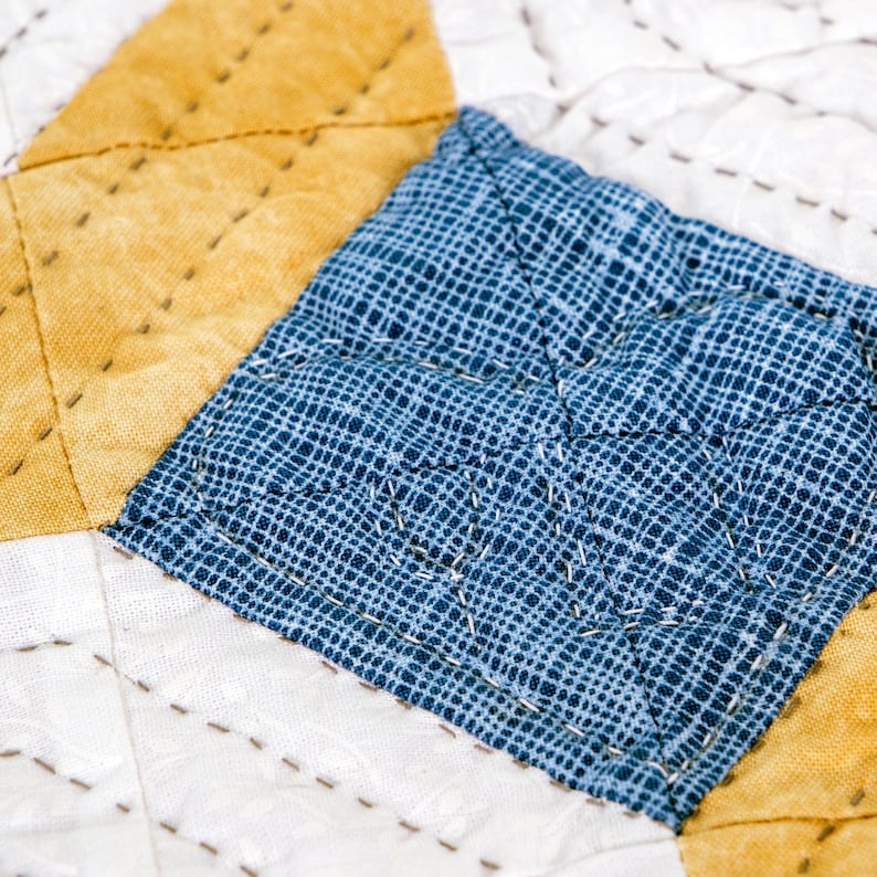 May include: Close-up of a handmade quilt with a patchwork design. The quilt features a blue and white patterned square, a yellow triangle, and a white triangle. The pieces are stitched together with white thread, creating a textured surface.