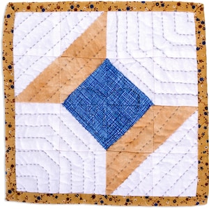 May include: Square quilted coaster with a central blue diamond, surrounded by tan and white geometric shapes. The edges are bordered with a floral patterned fabric in shades of brown, blue, and yellow. The stitching is visible.