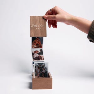 May include: A wooden box with a pull-out accordion photo display. The top wooden lid features the text Sally & Onur with small heart and infinity symbols. Three square photographs are attached in a vertical strip, showing people outdoors.