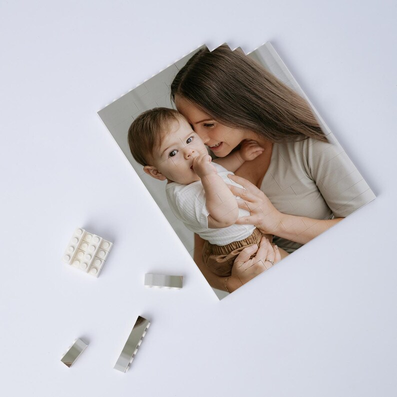 May include: A photograph of a mother holding a baby, with a white background. The baby is wearing a white shirt and brown shorts. Several white and silver toy blocks are scattered around the photo.