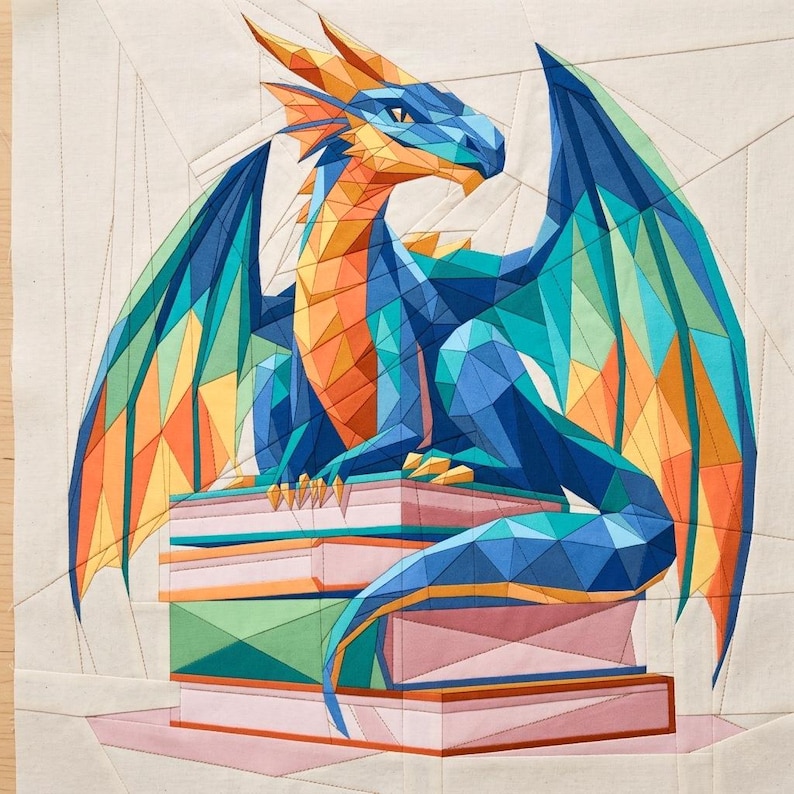 May include: A fabric quilt block featuring a geometric, low-poly style dragon in shades of blue, orange, and teal, perched atop a stack of books. The dragon has large wings and sharp angles, with visible stitching lines throughout the design.