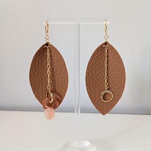 Könnte beinhalten: Ein Paar Ohrhänger mit großen, braunen, blattförmigen Lederanhängern. Jeder Ohrring hat eine goldfarbene Kette und eine kleine, rosa, durchscheinende Perle oder einen Ring. Die Ohrringe werden auf einem transparenten Ständer vor weißem Hintergrund präsentiert.