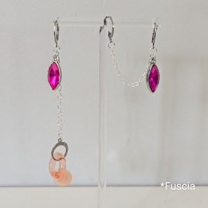 May include: A pair of asymmetrical earrings. One earring features a fuchsia-colored marquise-shaped gem, a silver chain, and two peach-colored circular charms. The other earring has a fuchsia-colored marquise-shaped gem. The word "*Fucsia" is in the image.