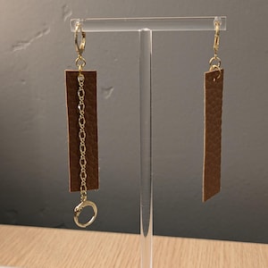May include: A pair of brown leather earrings with gold-coloured accents. One earring features a rectangular leather piece connected to a gold chain and a small gold hoop. The other earring is a simple rectangular leather piece.