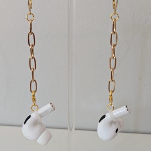 May include: A pair of white wireless earbuds with black accents, suspended from gold-colored chain earrings. The earrings are hanging against a white background.