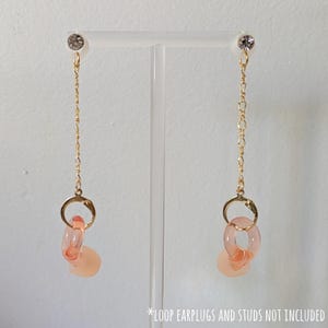 May include: A pair of dangling earrings with a gold chain and a clear crystal stud. The earrings feature a gold ring with two translucent pink circles and a pink teardrop-shaped charm. The earrings are displayed on a clear stand.