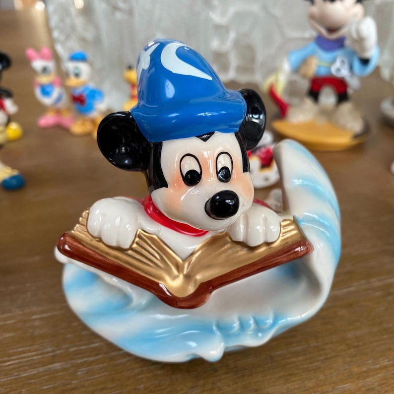 Op de afbeelding: Een keramieken beeldje van Mickey Mouse als tovenaar, met een blauwe hoed en witte maan. Mickey houdt een gouden boek vast en staat op een blauw-witte golfvormige basis. Andere Disney-beeldjes zijn op de achtergrond te zien.