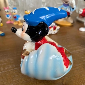 Op de afbeelding: Een keramieken beeldje van Mickey Mouse als de Tovenaarsleerling, met een blauwe hoed met witte accenten en een rode jurk. Het beeldje staat op een blauwe wolkenbasis, met andere Disney-beeldjes op de achtergrond.