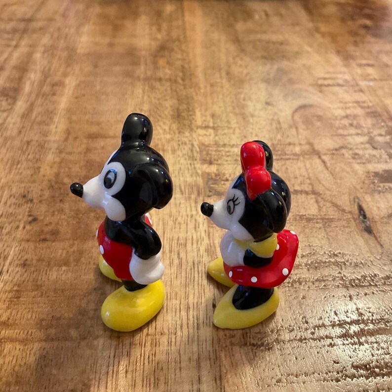 Op de afbeelding: Twee kleine beeldjes van Mickey en Minnie Mouse. Mickey draagt een rode korte broek, witte handschoenen en gele schoenen. Minnie draagt een rode polkadot rok, witte handschoenen, een rode strik en gele schoenen. Beide figuren hebben zwarte oren en staan op een houten oppervlak.