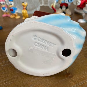 Op de afbeelding: Een keramieken beeldje met een blauw en wit ontwerp. De basis van het beeldje is wit en heeft de tekst "&copy; DISNEY CHINA". Andere Disney-beeldjes zijn zichtbaar op de achtergrond.