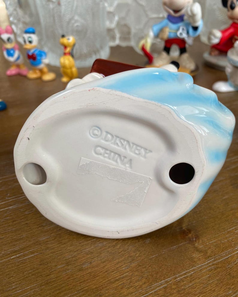 Op de afbeelding: Een keramieken beeldje met een blauw en wit ontwerp. De basis van het beeldje is wit en heeft de tekst "&copy; DISNEY CHINA". Andere Disney-beeldjes zijn zichtbaar op de achtergrond.