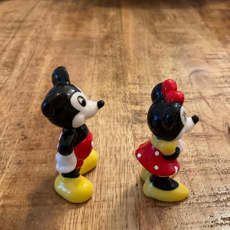 Op de afbeelding: Twee keramische beeldjes van Mickey en Minnie Mouse. Mickey draagt een rood en zwart outfit met gele schoenen. Minnie draagt een rode polkadot jurk met een rode strik en gele schoenen. Beide figuren hebben zwarte oren en witte gezichten.