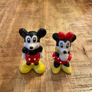 Op de afbeelding: Twee keramische beeldjes van Mickey en Minnie Mouse. Mickey draagt een zwart, rood, wit en geel outfit. Minnie draagt een rode jurk met witte stippen, een rode strik en gele schoenen. Beide hebben zwarte oren en witte gezichten.