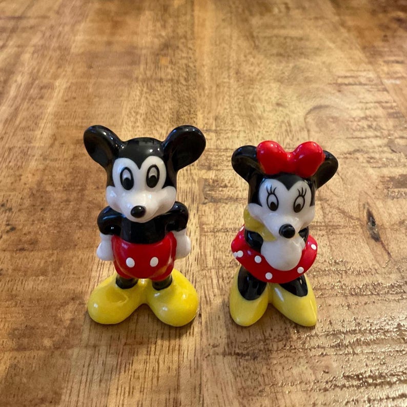 Op de afbeelding: Twee keramische beeldjes van Mickey en Minnie Mouse. Mickey draagt een zwart, rood, wit en geel outfit. Minnie draagt een rode jurk met witte stippen, een rode strik en gele schoenen. Beide hebben zwarte oren en witte gezichten.