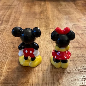 Op de afbeelding: Twee keramische beeldjes van Mickey en Minnie Mouse. Mickey draagt een rode broek, witte handschoenen en gele schoenen. Minnie heeft een rode polkadotjurk, een rode strik en gele schoenen. Beide hebben zwarte oren.