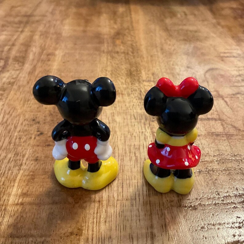 Op de afbeelding: Twee keramische beeldjes van Mickey en Minnie Mouse. Mickey draagt een rode broek, witte handschoenen en gele schoenen. Minnie heeft een rode polkadotjurk, een rode strik en gele schoenen. Beide hebben zwarte oren.