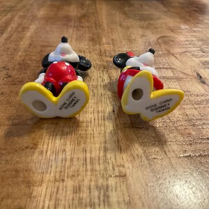 Op de afbeelding: Twee kleine Mickey Mouse beeldjes. De beeldjes staan in een klassieke pose, met zwarte oren, witte handschoenen, een rode broek en gele schoenen. De onderkant van elk beeldje leest "Bone China Disney Taiwan."