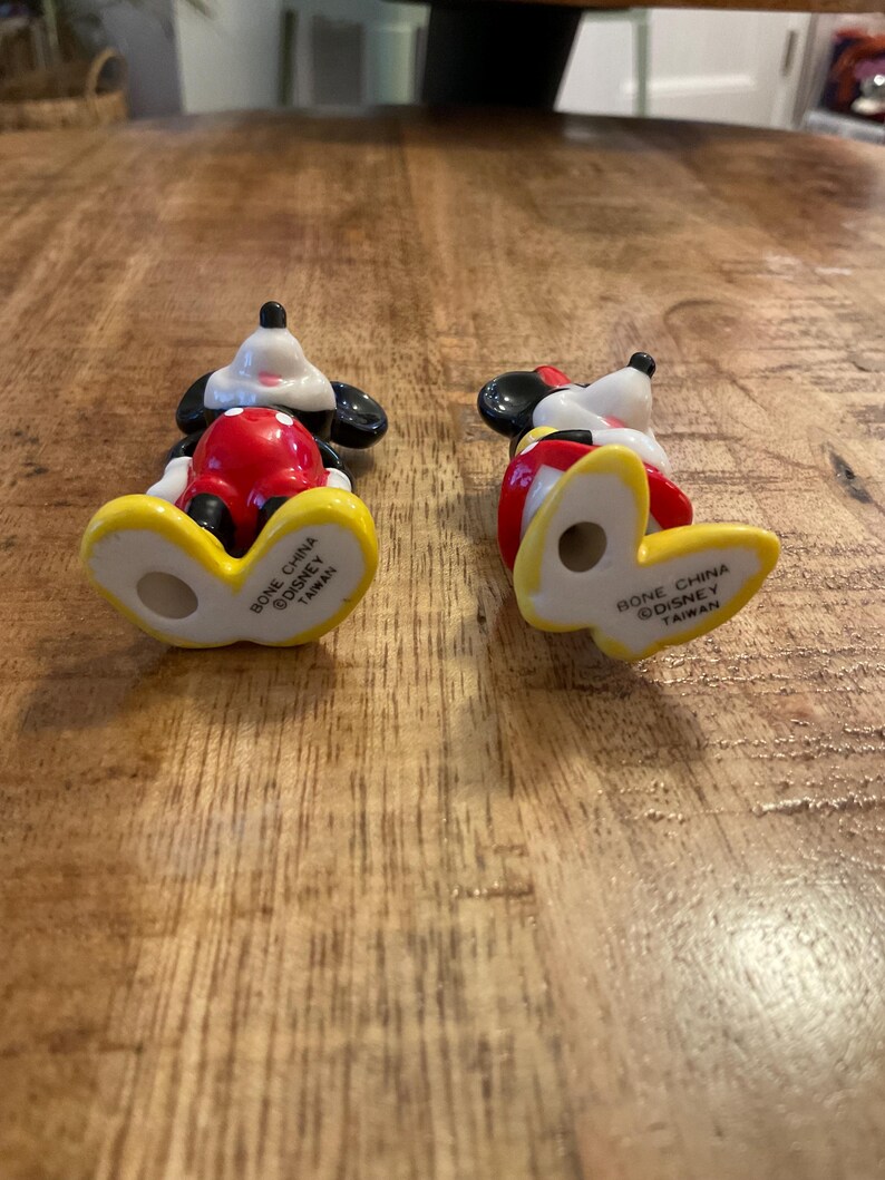 Op de afbeelding: Twee kleine Mickey Mouse beeldjes. De beeldjes staan in een klassieke pose, met zwarte oren, witte handschoenen, een rode broek en gele schoenen. De onderkant van elk beeldje leest "Bone China Disney Taiwan."