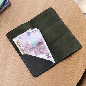 Può includere: Un portafoglio in pelle verde oliva scuro con una singola tasca diagonale. Il portafoglio contiene una banconota da 50 grivnie ucraine con il ritratto di Mykhailo Hrushevskyi visibile. Il portafoglio presenta cuciture visibili lungo i bordi.