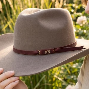 Könnte beinhalten: Ein taupefarbener Filzhut im Fedora-Stil mit breiter Krempe und einem dunkelroten Band. Das Band ist mit einer kleinen, rechteckigen, goldfarbenen Metallplakette verziert, in die die Großbuchstaben AB eingraviert sind.
