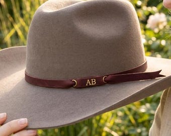 May include: A taupe felt fedora hat featuring a wide brim and a deep burgundy ribbon band. The ribbon band is accented with a small, rectangular gold-toned metal plaque engraved with the capital letters AB, positioned at the front of the hat.