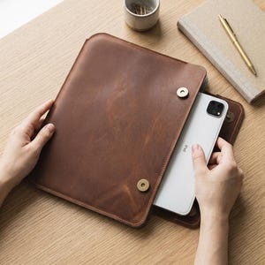 Peut inclure: Une housse pour tablette en cuir marron dotée d'un rabat fermé par deux boutons-pression métalliques de couleur laiton. Une tablette blanche est partiellement insérée dans la housse, révélant une partie de son boîtier et de son objectif.