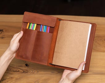 May include: Open brown leather notebook with a flap closure and two pockets filled with colorful pens. The notebook is open, revealing a stack of blank, cream-colored paper. The leather has a rich, warm tone, and the stitching is visible.