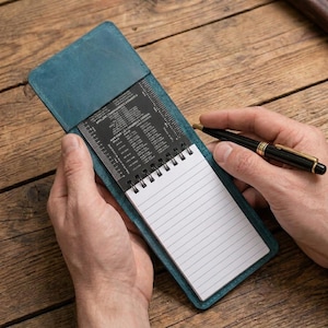 Puede incluir: Una funda de cuero azul cerceta para cuaderno que contiene un bloc de notas en espiral con papel blanco rayado. Una tarjeta de referencia de metal negro con datos técnicos y escalas de medición está fijada en la parte superior. Un bolígrafo negro.