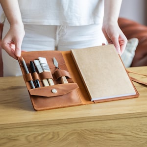 Puede incluir: Un organizador de suministros de arte de cuero marrón con un cuaderno y varias herramientas de arte. El organizador contiene un lápiz, bolígrafos, pinceles y una espátula. El cuaderno tiene una cubierta marrón y páginas en blanco.