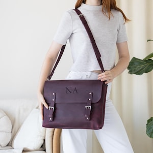 May include: A burgundy leather messenger bag with a flap closure and adjustable shoulder strap. The bag features silver-colored buckles and the initials "NA" embossed on the front. The bag is held by a person wearing a gray shirt and white pants.