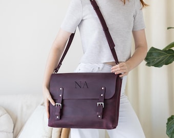 May include: A burgundy leather messenger bag with a flap closure and adjustable shoulder strap. The bag features silver-colored buckles and the initials "NA" embossed on the front. The bag is held by a person wearing a gray shirt and white pants.