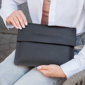 Könnte beinhalten: Eine dunkelgraue Laptop-Hülle aus Leder mit Klappenverschluss. Die Hülle hat ein minimalistisches Design mit einer Nahtdetail oben. Das Produkt wird von einer Person gehalten, die ein weißes Hemd und blaue Jeans trägt.