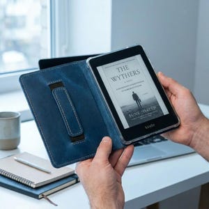 Puede incluir: Una funda de cuero azul para Kindle con un lector de libros electrónicos Kindle negro. El lector muestra la portada del libro "The Withers" de Fune Shapeh. La funda tiene una correa de mano y costuras en contraste.
