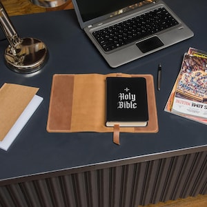 May include: A black Holy Bible with white gothic lettering and a cross, inside a tan leather book cover. The cover is open on a dark desk, next to a laptop, pen, and magazine.
