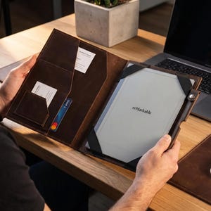 May include: A dark brown leather folio case holding a gray reMarkable tablet secured by black elastic corner straps. The interior features multiple slots for business cards and a credit card, with a metal snap closure on the right side edge.