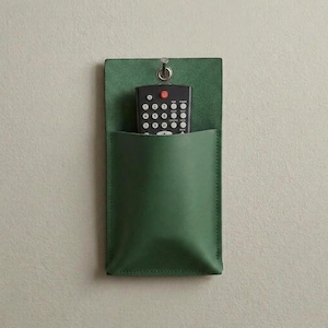 Peut inclure: Un organisateur mural en cuir vert forêt doté d'un panneau arrière rectangulaire avec un œillet métallique en haut pour la suspension. Une télécommande noire avec des boutons blancs est insérée dans la pochette de rangement profonde.
