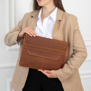 May include: A brown leather laptop sleeve with a flap closure. The sleeve is held by a person wearing a tan blazer and a white shirt. The leather has a textured appearance.