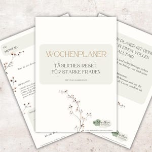 Könnte beinhalten: Drei weiße Papierplaner mit floralen Akzenten. Der zentrale Planer trägt den Text "WOCHENPLANER TÄGLICHES RESET FÜR STARKE FRAUEN". Die anderen Planer haben Text auf Deutsch, darunter "TAG MONTAG".