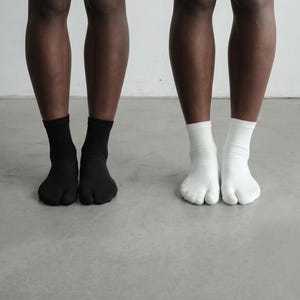 May include: A pair of black split-toe socks and a pair of white split-toe socks. These ankle-length socks feature a distinct separation between the big toe and the remaining toes, designed to be worn with traditional footwear or sandals.