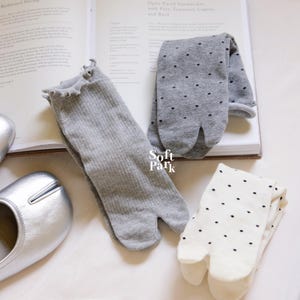May include: Three pairs of tabi socks in the image. One pair is gray with a ruffled top, another is gray with black polka dots, and the third is white with black polka dots. The socks are displayed on a book with text and a white surface.