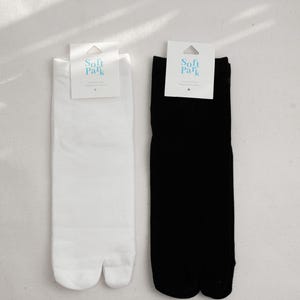 May include: Two pairs of tabi socks, one white and one black, displayed side-by-side. Each sock has a split toe design and a tag that reads "Soft Park". The socks are made of a soft material and are designed for comfort.