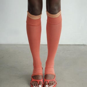 Puede incluir: Un par de calcetines hasta la rodilla en un tono naranja coral apagado con un puño acanalado de color tostado claro en la parte superior. Los calcetines presentan un diseño de punta abierta, dejando los dedos al descubierto, y se combinan con sandalias de tiras a juego.