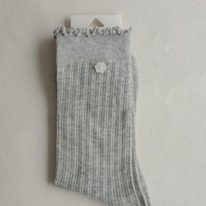 May include: A single gray ribbed sock with a white flower-shaped button. The top edge has a decorative, ruffled trim. The sock is displayed on a white background, attached to a white product tag.