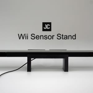May include: Black Wii sensor stand with the text "Wii Sensor Stand". The stand is rectangular with a central support and a power cord. The stand is designed to hold the Wii sensor bar.