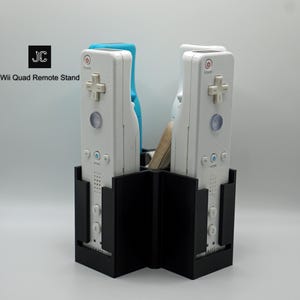 May include: Black Wii Quad Remote Stand holding four white and blue Wii remotes. The stand has a unique design with the text "Wii Quad Remote Stand" and the initials "JC" in a black square.