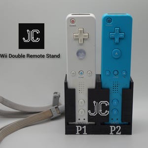 May include: A black stand holding two Wii remotes, one white and one blue. The stand has the text "JC" and "P1" and "P2". The text "Wii Double Remote Stand" is also visible. A gray wrist strap is in the foreground.