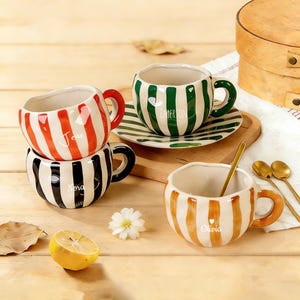May include: A set of ceramic teacups with striped patterns in red, black, green, and orange. Each cup features a unique design, with names such as "Jesse", "Mora", "Emerson", and "Olivia". Gold spoons and a lemon half are also visible.