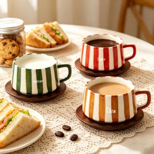 May include: Three striped ceramic mugs on wooden coasters. One mug has green and white stripes and the name "Adrian". Another has red and white stripes with the name "Claire". The third has brown and white stripes and the name "Eliza".