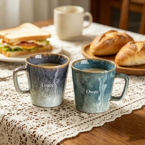 May include: Two ceramic mugs with handles, one blue and one teal, each with a name printed on the side. The mugs are filled with a beverage and sit on a lace tablecloth. A sandwich and bread rolls are also visible.