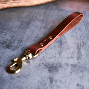 Op de afbeelding: Een bruin leren sleutelhanger met een messing sluiting en klinknagels. De leren lus is bevestigd aan een goudkleurige metalen sluiting. De sleutelhanger is ontworpen voor het vasthouden van sleutels of het bevestigen aan een tas.